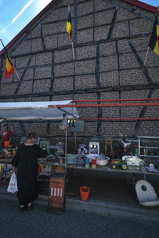 Dit is een foto van de luikse markt in Wahlwiller. Er staat een huis op  met daarvoor een marktkraam met diverse spullen. Enkele mensen kijken er geïnteresseerd naar. 